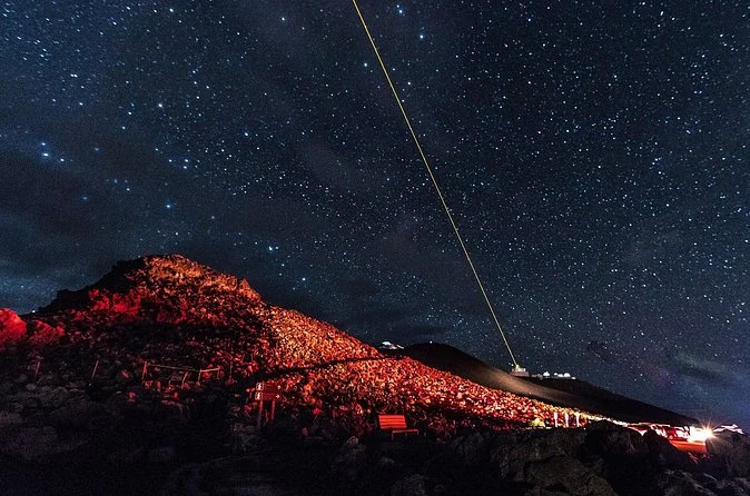 A starry night sky over the dark summit, a thin beam of light rising from the observatory area and a red glow on the slope