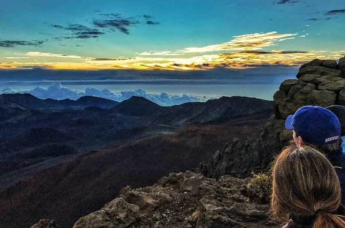 A visitor in a cap seen from behind looking over dark ridges at a pale dawn sky