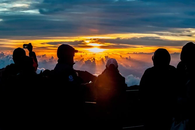 Dark silhouettes of people holding up phones against a sunrise breaking through cloud