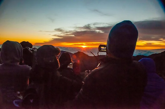 Hooded figures in silhouette watching an orange sunrise over the clouds