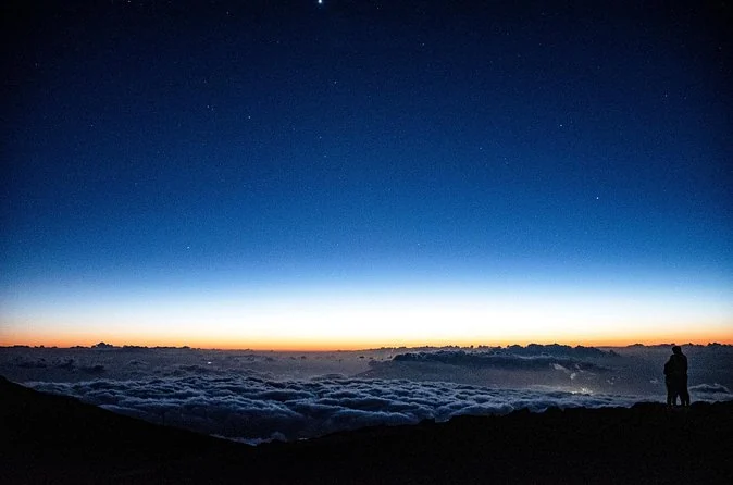 A deep blue dusk sky over a band of orange on the horizon above the clouds, with a lone figure on a ridge